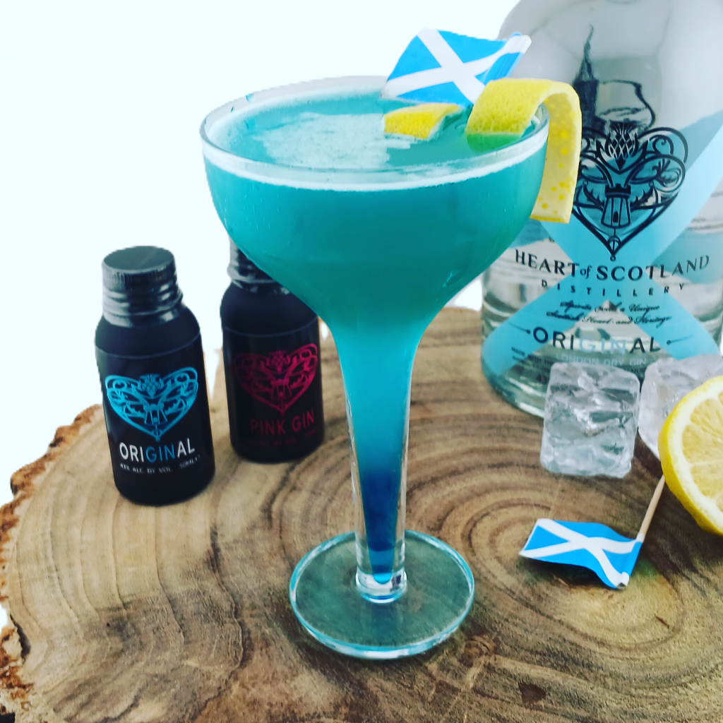 The Saltire cocktail in a coupe glass, layered with a luminous blue base and topped with a frothy white layer, garnished with lemon peel