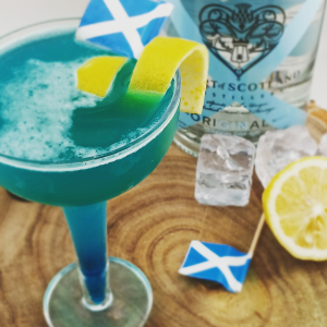 The Saltire cocktail in a coupe glass, layered with a luminous blue base and topped with a frothy white layer, garnished with lemon peel