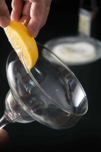 Hand running a lemon wedge around the rim of a cocktail glass before dipping into rimming sugar